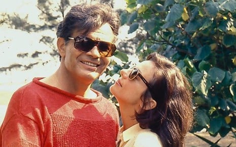 Os atores Tarcísio Meira e Bruna Lombardi estão abraçados e de óculos escuros como os personagens Renato Villar e Lúcia Brandão da novela Roda de Fogo (1986)