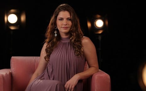 Com um vestido roxo, Ana Paula Tabalipa está sentada em um sofá avermelhado e olha para a câmera com expressão séria