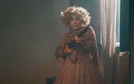 A atriz Paula Cohen como Lota, com uma peruca loira, cacheada e desgrenhada, segura uma espingarda com as duas mãos e com cara de assustada em cena de Nos Tempos do Imperador