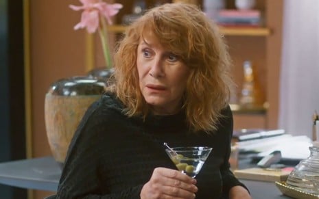 Renata Sorrah caracterizada como Wilma; ele tem os cabelos ruivos na altura dos ombros, usa uma blusa branca e segura uma taça de martini em cena de Vai na Fé