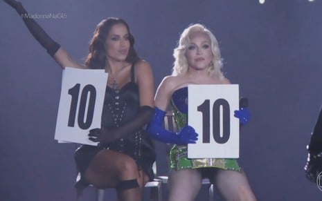 Anitta e Madonna estão sentadas em banquinhos e seguram placas com a nota 10