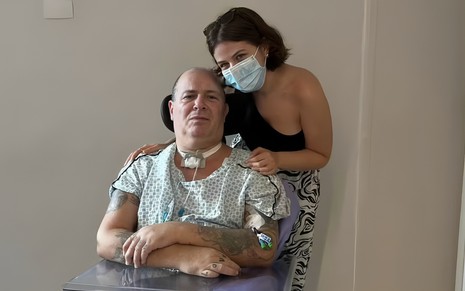 Isabella Aglio posa com o pai, o baixista Mingau, no hospital; ele usa trajes hospitalares, e ela uma máscara