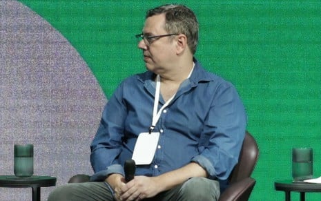 Com blusa azul e calça cinza, João Emanuel Carneiro está sentado, segurando um microfone, e olhando para o lado no palco do Festival Acontece