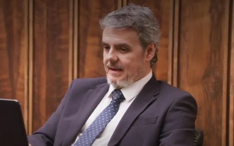 O ator Cassio Gabus Mendes em cena como Roberto em Elas Por Elas, de terno e gravata, expressão séria
