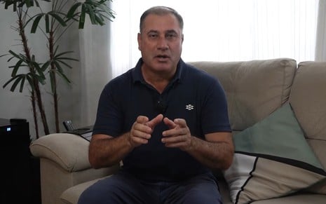 O jornalista César Galvão em reportagem para seu canal no YouTube; ele está em um sofá e veste camisa preta